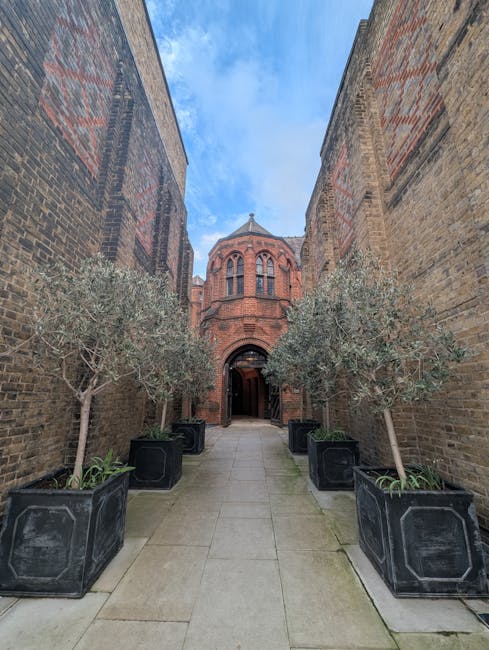 A narrow outdoor corridor surrounded by tall brick walls leading to an ornate red brick building with arched windows and a domed roof. On either side of the paved pathway, there are several large blac