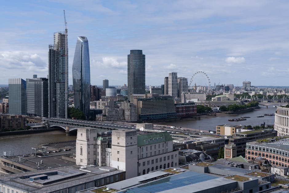 A sweeping aerial view of the London skyline featuring skyscrapers such as the twisting shard-shaped building and other high-rise office towers, with the River Thames winding through the city below. I